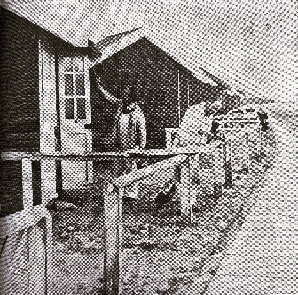 Black-and-white photo showing painters at work painting the vintage St Annes Beach Huts along the seaside